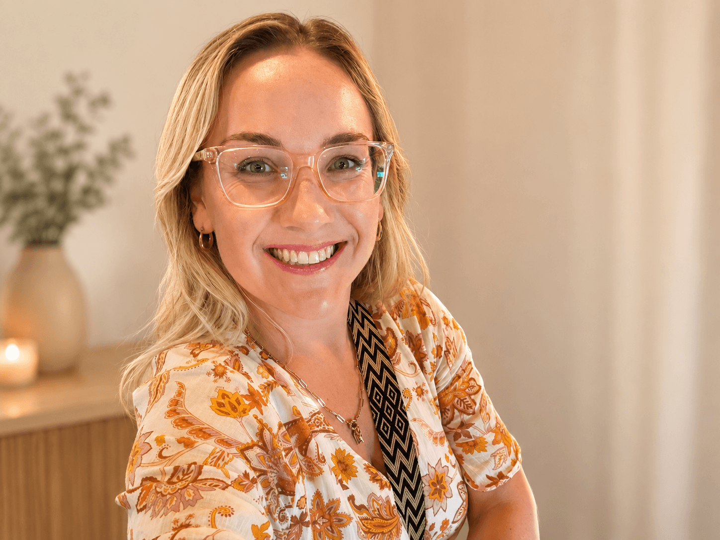 Smiling blonde woman wearing clear-framed glasses and a floral print blouse in warm lighting.