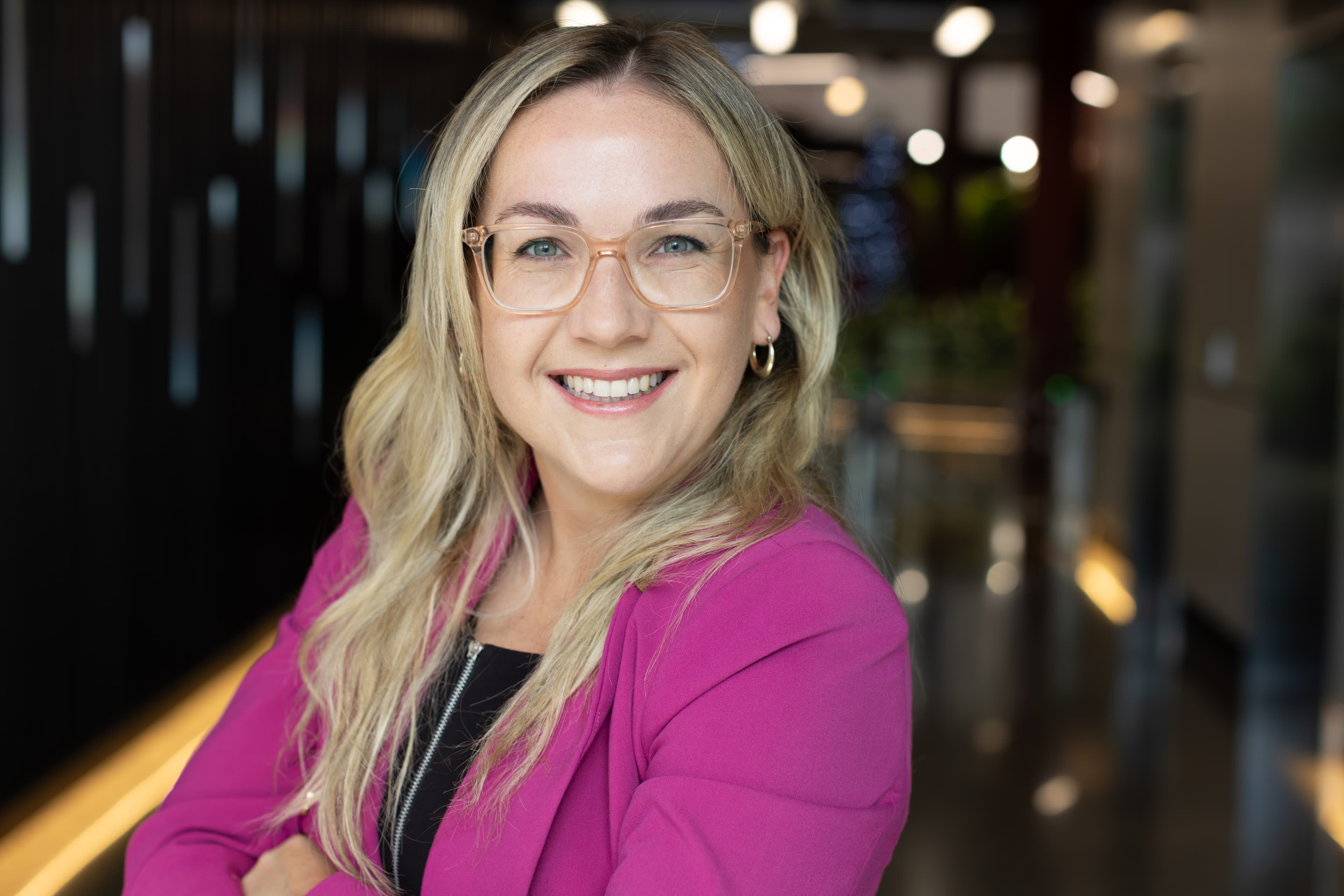 Smiling blonde woman wearing clear glasses and a bright pink blazer in a modern office.